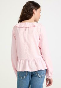 Ung kvinde med langt brunt hår har en lys pink langærmet bluse med flæsekanter og teksturerede prikker, parret med blå jeans.