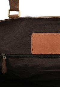 Wnętrze brązowej torby z płótna z kieszenią na zamek błyskawiczny i skórzanym akcentem. Zawiera prostokątną skórzaną naszywkę do identyfikacji. Teksturowana powierzchnia.