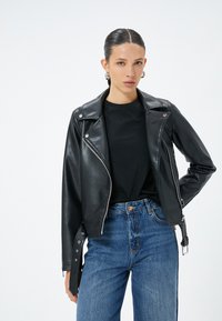 Svart läderbikerjacka med silverdetaljer, dragkedjefickor och knappkrage, bärs över en svart skjorta och blå högmidjade jeans.