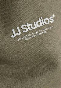 Ύφασμα ελιάς με λευκό τυπωμένο κείμενο "JJ Studios" και μικρότερο κείμενο από κάτω που λέει "Σας το φέρνει η ομάδα J&J, σχεδιασμένο στη Δανία."