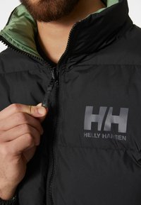 Zwarte pufferjack met een groene voering, ritsopening en een opvallend grijs Helly Hansen-logo op de borst. Gladde textuur en gewatteerd ontwerp.