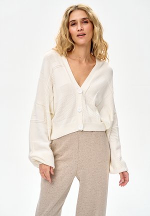 Kobieta o falujących blond włosach, ubrana w biały, teksturowany, zapinany na guziki kardigan oraz beżowe spodnie z wysokim stanem na jednolitym białym tle.