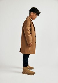 Pruun villane mantel mustade nuppudega, seljas tumesinine pükste ja tan värvi saapadega. Subjekt seisab külg ees, demonstreerides mantli pikkust ja tekstuuri.