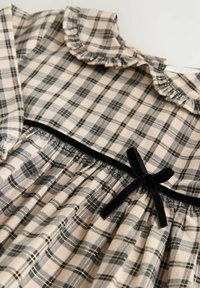 Kleid mit Karomuster, das einen voluminösen Halsausschnitt, eine geraffte Taille und eine schwarze Samtschleife als Akzent aufweist. Das Material ist aus weicher Baumwolle mit beigen, blauen und goldenen Farbtönen.