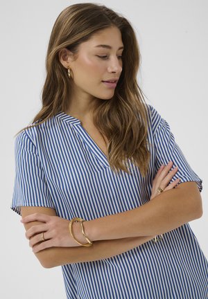 Žena s dlhými hnedými vlasmi, ktorá má na sebe modrobielo pruhované tričko s krátkym rukávom, zlaté kruhové náušnice, náramky a prstene, pozerajúca sa do strany.