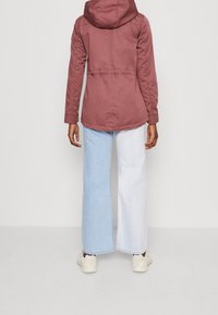 Lehká bunda s kapucí v barvě prachové růže, s přitažením v pase a mírně zaobleným lemem. Kombinována s světle modrými džínami s širokými nohavicemi a teniskami.