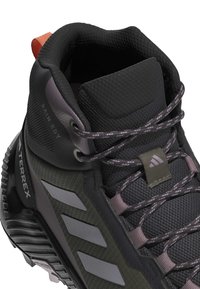 Scarpa da trekking a metà altezza con tomaia sintetica nera, pannelli in rete, accenti grigi e lacci testurizzati. Include un anello di estrazione e una suola decorata.