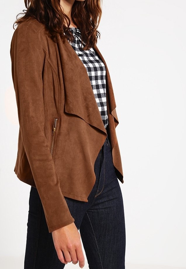 Femme portant une veste en daim marron par-dessus une chemise à carreaux noire et blanche et un jean bleu foncé, debout devant un fond blanc.