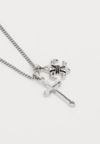 Sølvkjede med to pendanter: et detaljert kors og en stilisert fleur-de-lis med svarte aksenter.