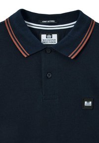 Jūrinės spalvos polo marškiniai su rudomis dvigubomis juostelėmis ant apykaklės, dviem mygtukais ir "Weekend Offender" logotipo užleidimo pleistru ant krūtinės bei apykaklės etiketėje.