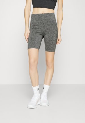Donna con pantaloncini da ciclista grigi a vita alta, top corto nero, calzini bianchi Adidas e sneakers bianche, in piedi su sfondo chiaro e uniforme.
