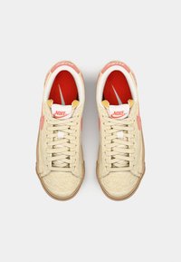 Beige suede sneakers with red accents, white laces, and padded interior. Features a prominent Nike logo and gum rubber sole.