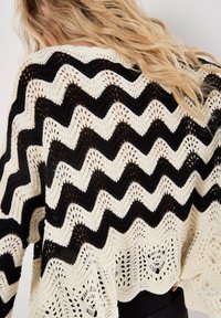 Schwarzer und cremiger gestrickter Poncho mit einem Zickzackmuster, das über intricate offene Details und strukturierten Stoff verfügt, gestaltet für eine Übergröße.