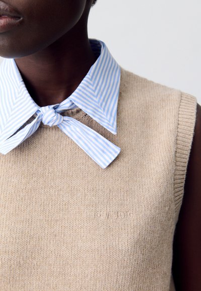 Maglione lavorato a maglia beige senza maniche con una finitura testurizzata, abbinato a una camicia a righe blu chiaro e bianca con colletto legata al collo.