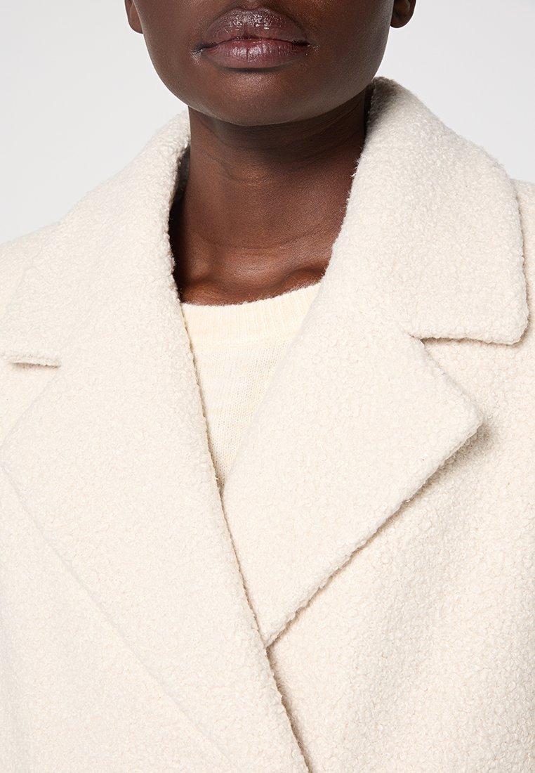 Osoba o ciemnej karnacji ubrana w kremowy, fakturowany wełniany płaszcz na jasnym swetrze, przycięta tak, aby pokazać usta, szyję i górną część klatki piersiowej.