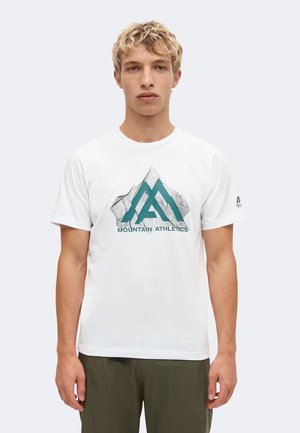 Ung mand med kort blondt hår iført en hvid t-shirt med et bjergmotiv og teksten "Mountain Athletics", stående mod en ensfarvet baggrund.