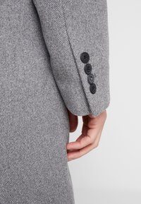 Manica di un cappotto grigio di lana con un tessuto testurizzato e quattro bottoni neri sul polsino, che mostra un primo piano del braccio.