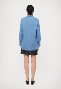 Hellblaues Jeanshemd mit langen Ärmeln und geknöpften Manschetten, das einen geschwungenen Saum hat. Getragen über einem engen schwarzen Lederrock.