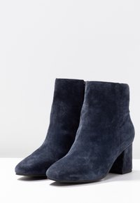 Botines de ante azul marino con punta redondeada, cierre de cremallera en la parte trasera y tacón grueso, que presentan una textura suave y un diseño elegante.