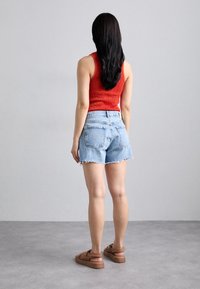 Woman with long black hair wearing a red sleeveless top, light blue denim shorts, and brown sandals stands facing away against a plain gray wall.