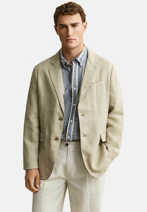 Jonge man die een beige blazer draagt over een blauw-wit gestreept overhemd en lichtbeige broek, staand met één hand in de zak.