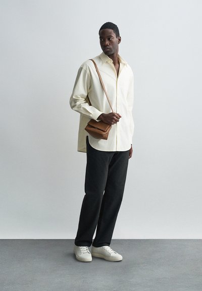 Jasno beżowa, długa koszula z luźnym krojem, zestawiona z ciemnymi spodniami i białymi sneakersami. Brązowa torba na ramię dodaje głębi.