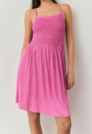 Femme portant une robe rose sans manches avec un corsage froncé et une jupe fluide, debout devant un fond clair uni.