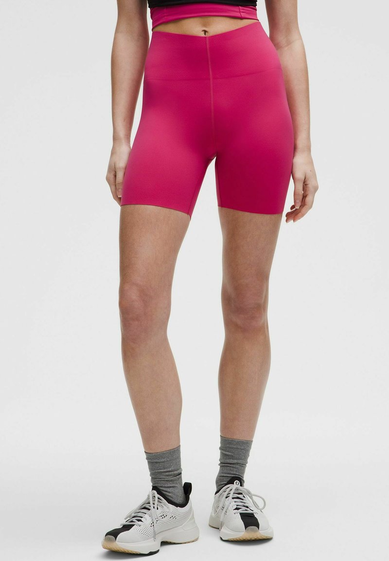Shorts da ciclismo rosa aderenti con vita alta, realizzati in materiale elastico. Indossati con calze grigio chiaro e scarpe sportive nere e bianche.