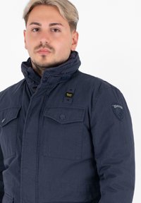 Giacca imbottita blu navy con colletto alto, due grandi tasche sul petto, chiusure a bottoni e velcro, e un patch del logo sulla spalla.