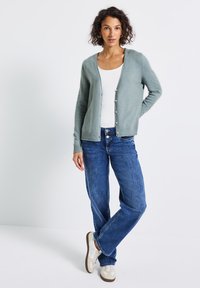 Cardigan vert clair en tissu doux, avec une fermeture à boutons, des poignets côtelés et une coupe décontractée. Assorti à un jean bleu et des baskets claires.