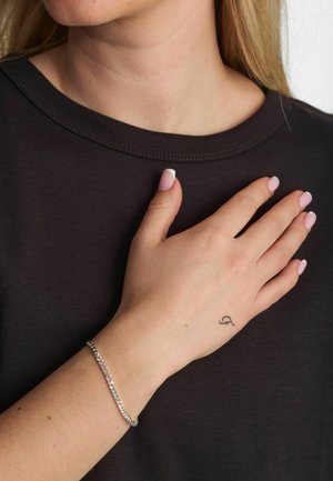 Czarna koszula z krótkim rękawem, zbliżenie na dłoń z srebrną bransoletką łańcuszkową i małym tatuażem na nadgarstku, jasnoróżowy lakier do paznokci.