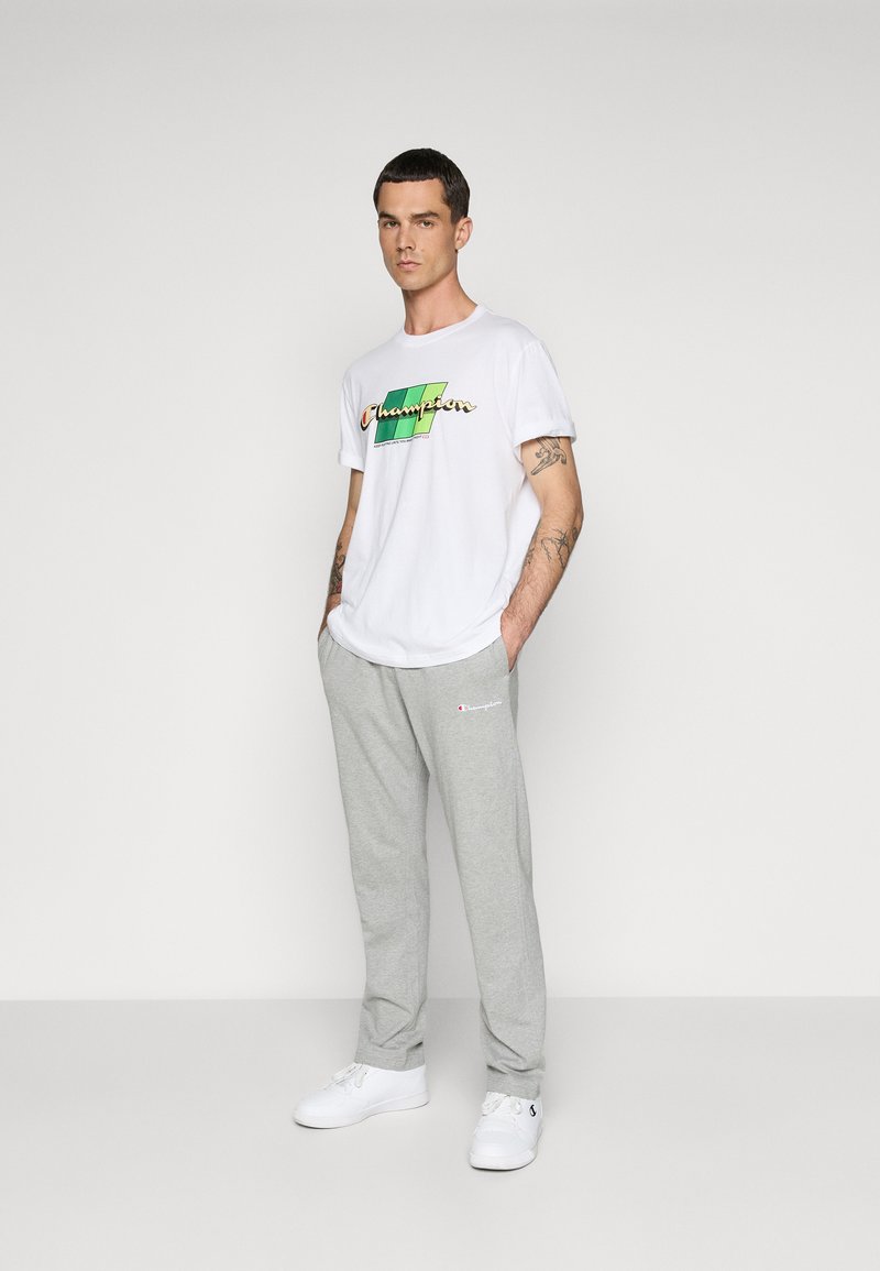 Camiseta blanca con el colorido logo de "Champion", combinada con pantalones de chándal grises claros que tienen un pequeño logo. El modelo lleva zapatillas blancas.