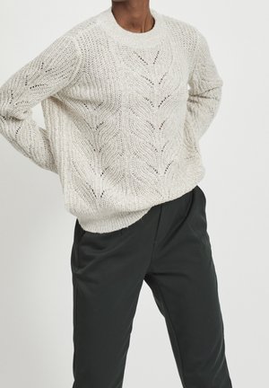 Personne portant un pull en maille crème avec des motifs de feuilles et un pantalon noir taille haute, debout les mains sur les hanches.