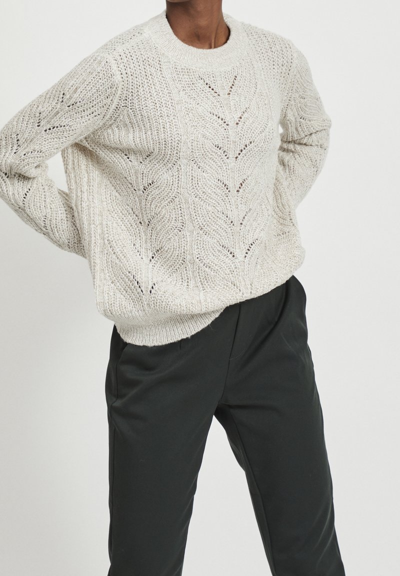 Personne portant un pull en maille crème avec des motifs de feuilles et un pantalon noir taille haute, debout les mains sur les hanches.