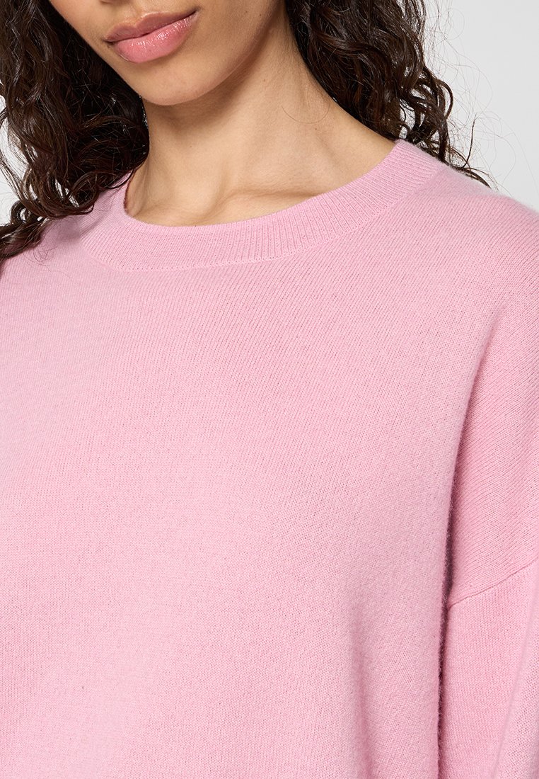 Jasnoróżowy sweter dzianinowy z okrągłym dekoltem i miękką teksturą. Projekt zawiera opadające ramiona, co zapewnia luźniejszy krój.