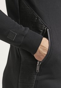 Felpa nera con pannello laterale testurizzato, tasca con zip, polsini a costine e una piccola etichetta con marchio sulla manica.
