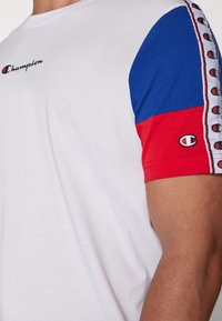 Camiseta blanca con mangas cortas azules y rojas, que presenta un logotipo en el pecho y una franja estampada a lo largo de los bordes de las mangas.