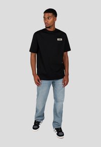 Czarna bawełniana koszulka z okrągłym dekoltem i krótkimi rękawami, z prostokątną białą metką z tekstem. Noszona z jasnoniebieskimi dżinsami i czarnymi trampkami.