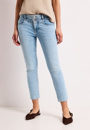 Kvinde iført lyseblå jeans med høj talje og dobbelt knaplukning samt brune spidse flats, stående mod en ensfarvet lys baggrund.