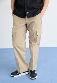 Beige cargo bukser med sidelommer, lige snit, lavet af bomuld, sammen med sorte sneakers med hvid sål og accenter.