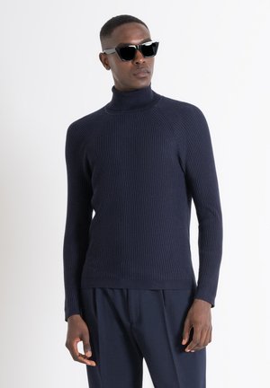 Suéter de canalé azul marino con cuello alto y mangas raglán, hecho de un tejido texturizado. Modelo combinado con gafas de sol negras.