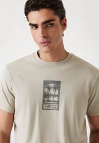 Beige T-shirt met korte mouwen met een zwarte grafische print van palmbomen en een landschap, met het label "GUESS AUTHENTIC DESIGN."
