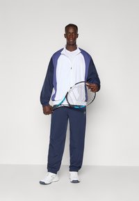 Atletska obleka, ki vključuje belo in mornarsko modro tekaško jakno, mornarsko hlače in bele športne copate. Oseba drži teniško lopar s modrim poudarkom.