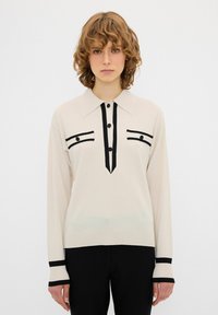 Maglione crema con accenti neri. Presenta un colletto, chiusura con bottoni e strisce verticali sul davanti. Maniche e orlo a coste.