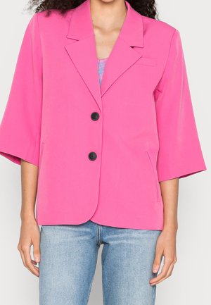 Kvinde iført en skarpt pink blazer med trekvartærmer og sorte knapper, kombineret med lyseblå jeans.