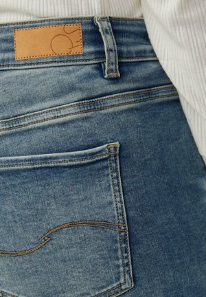 Verwaschene blaue Jeans aus Denim mit einem hellbraunen Lederpatch am Taillenbund, mit einer einzelnen Gesäßtasche mit geschwungenen Steppnähten.