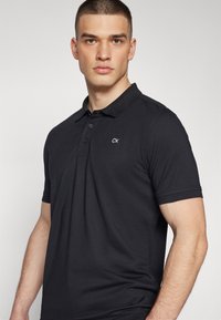 Crna polo majica izrađena od mješavine pamuka, s tri gumba na prednjem dijelu i CK logom na lijevoj strani grudi. Kratki rukavi i klasičan kroj.