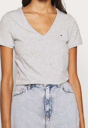 Kobieta w jasnoszarej koszulce z krótkim rękawem i dekoltem w serek, z małym czerwono-biało-niebieskim logo na lewej piersi, włożona w jasnoniebieskie dżinsy z wysokim stanem.