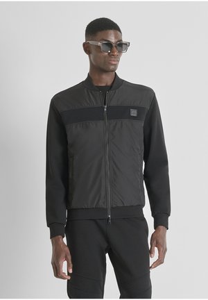 Black bomber jacket made of quilted fabric with ribbed cuffs and a front zip. Features a logo patch and paired with black pants.