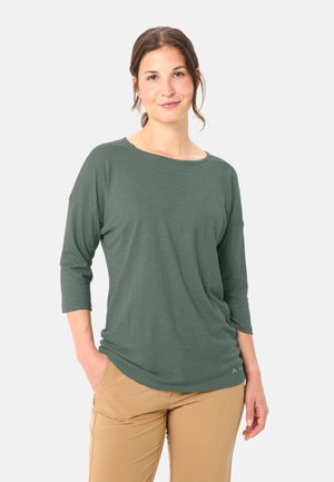 Femme aux cheveux bruns attachés en queue de cheval basse, portant une chemise verte à manches longues et un pantalon beige, debout avec une main dans la poche.